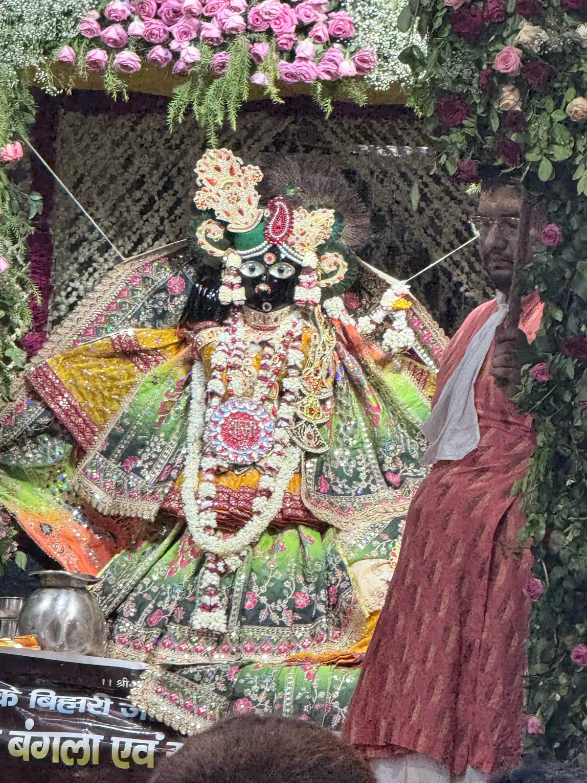 Thakur Banke Bihari ji in a green and gold poshak at Banke Bihari Temple, Vrindavan, beneath a flower canopy, with a Hindi banner in front of the shrine