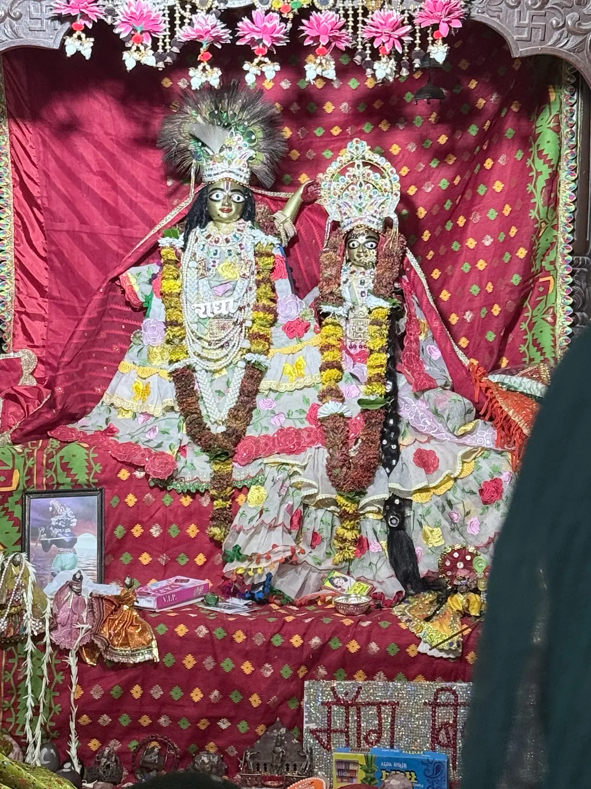 Radha Krishna deities side by side as bride and groom in white floral poshak with peacock feather crowns at the temple in Bhandir Van