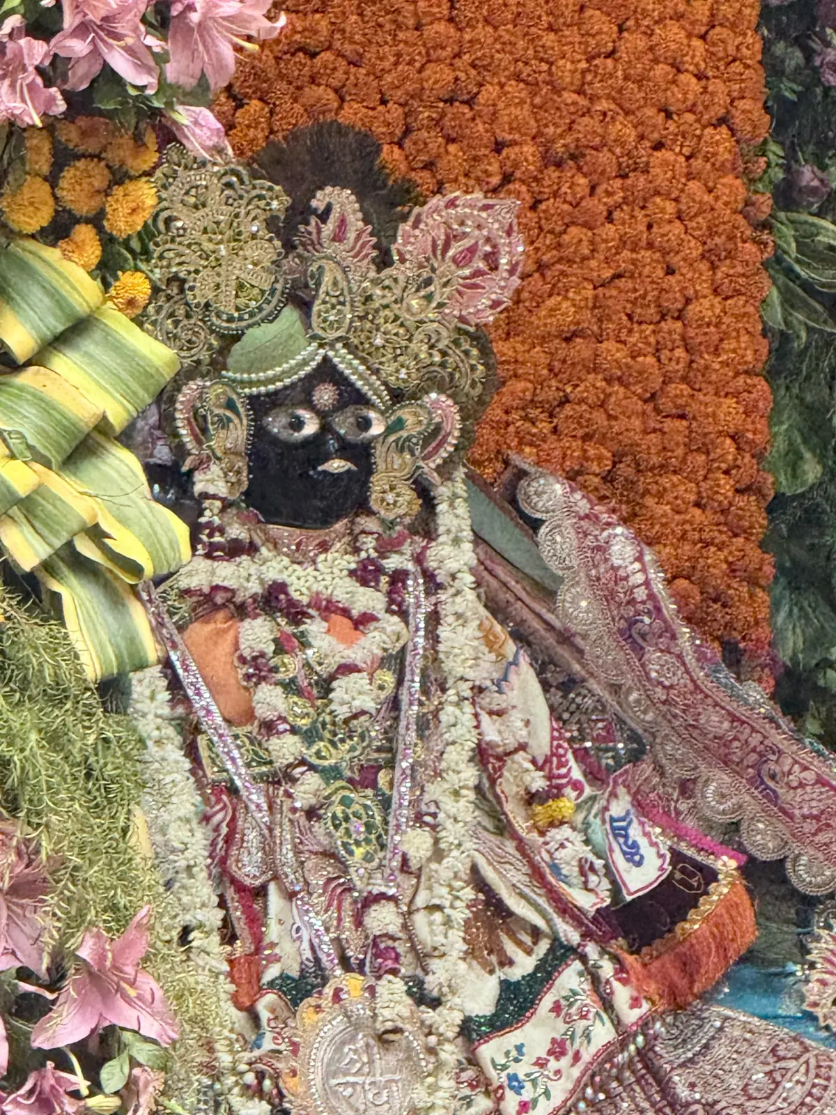 Black stone deity of Lord Krishna in a richly embroidered poshak with a jewelled crown and white and pink garlands before a marigold backdrop