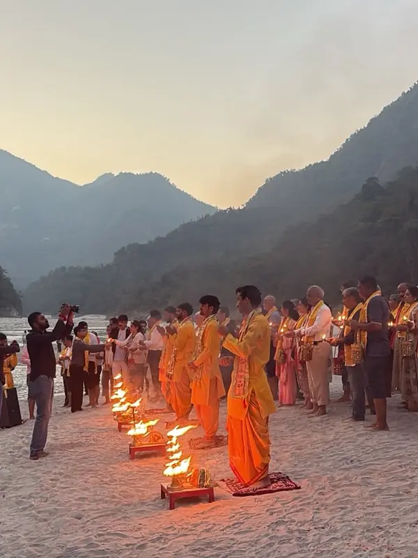 House of Ullassa group performing a private evening Ganga Aarti on a sandy ghat by the river in Rishikesh, with hills behind