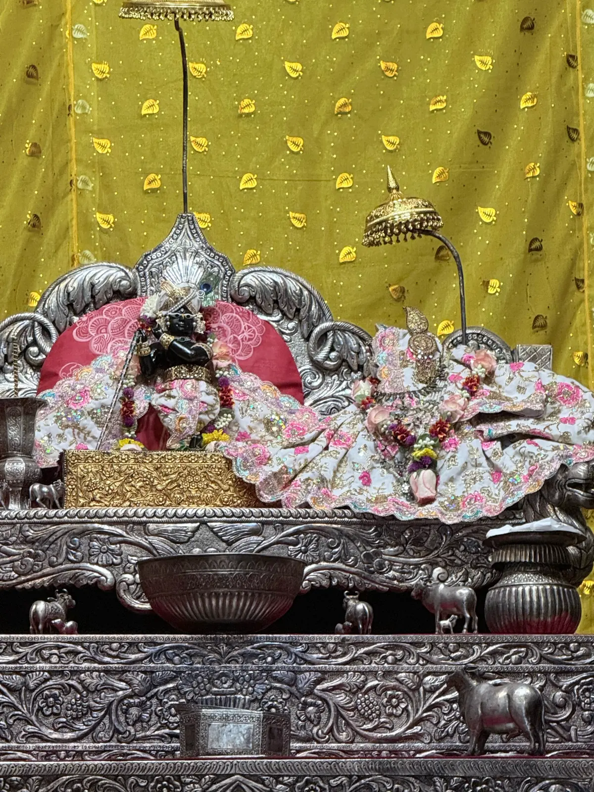 The silver altar at Sri Radha Raman Temple, Vrindavan, with the black deity of Sri Radha Raman holding a flute beside Radha's gaddi in pink and white poshak